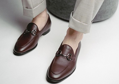 A person is wearing brown leather loafers with metallic horsebit details and light-colored pants.