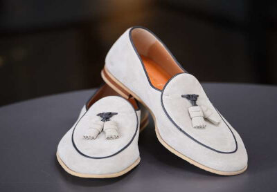 A pair of light gray suede loafers with tassels and dark piping, displayed on a dark surface.