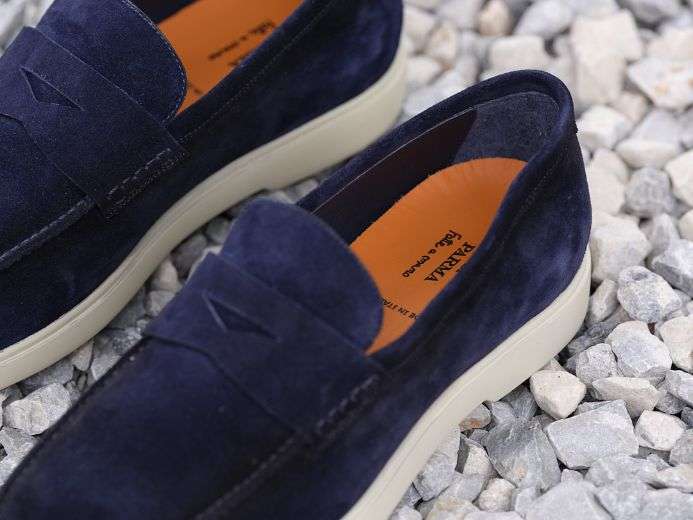 A pair of navy blue suede loafers with white soles, placed on a bed of gray gravel.