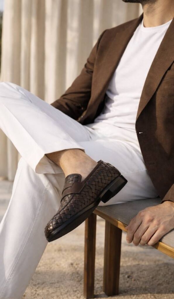Un uomo che indossa un blazer marrone, pantaloni bianchi e scarpe uomo fatte a mano siede su una panchina di legno con le gambe incrociate.