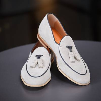 Un paio di mocassini in pelle scamosciata grigio chiaro con finiture e nappe nere, queste scarpe uomo fatte a mano sono elegantemente esposte su una superficie scura.