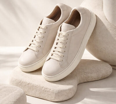 A pair of beige lace-up sneakers displayed on abstract, light-colored stone shapes with soft shadows in the background.